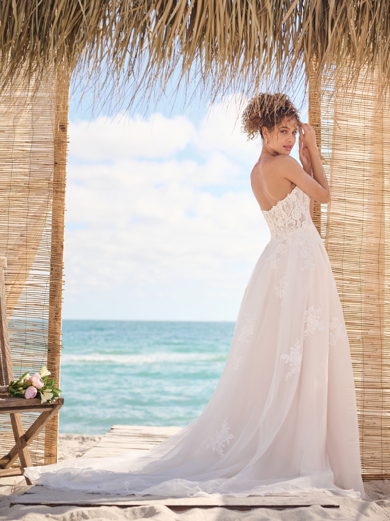 A bride poses under a thatched canopy in a flowing strapless gown with lace detail and a long train, framed by the turquoise waves.