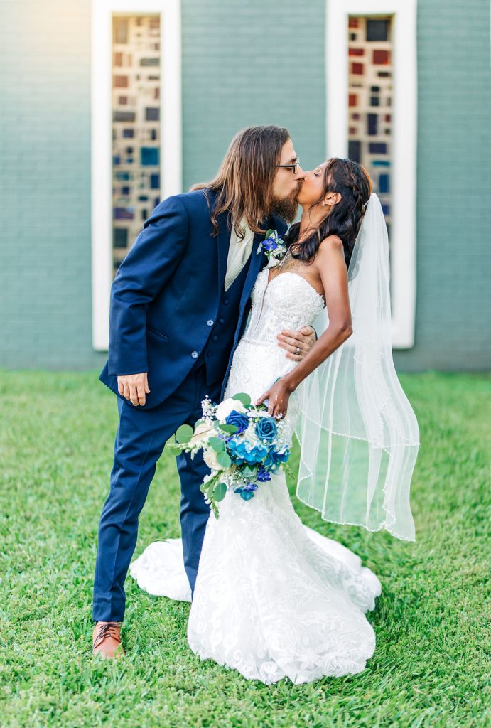 Groom kisses bride in a strapless wedding dress as they pose on green grass with stained glass windows behind them.