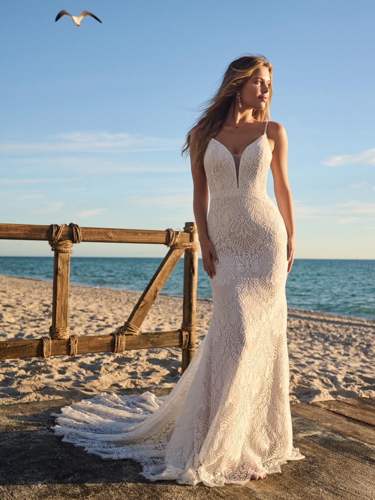A bride stands barefoot on the sand in a figure-hugging lace gown with a dramatic train and plunging neckline, perfect for a beachside wedding.