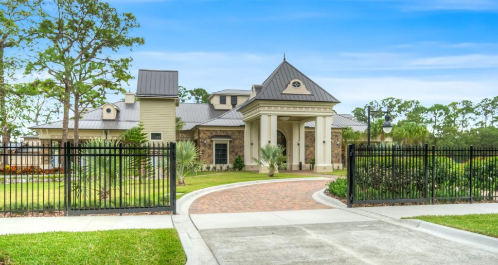 Grand building with a gated entrance, brick driveway, and manicured landscaping, set against a bright blue sky.