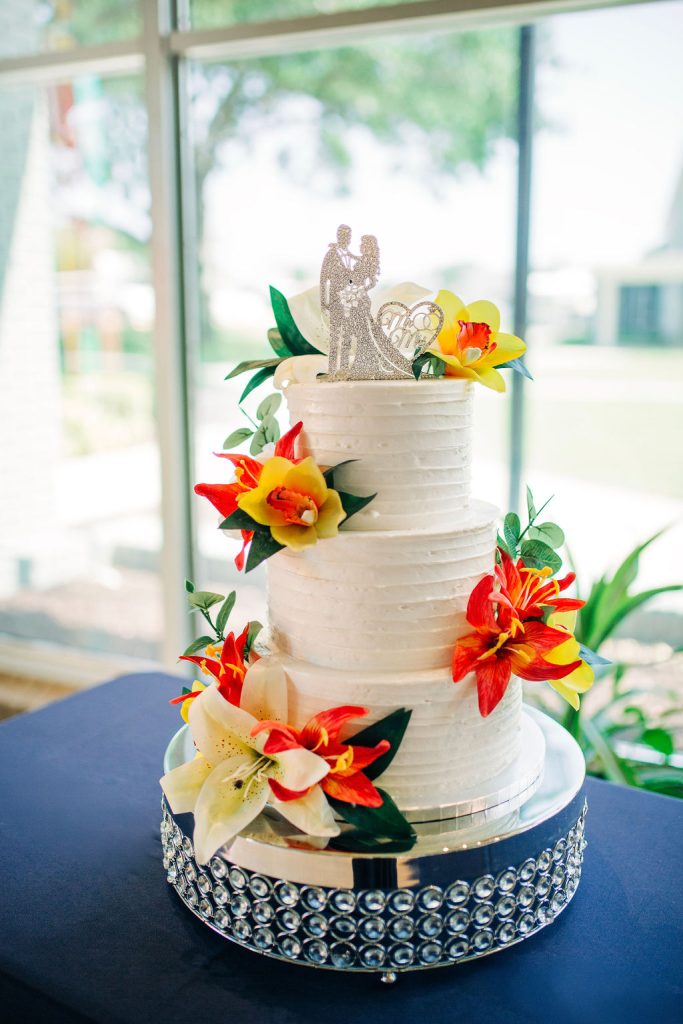 White wedding cake with bright red and yellow flowers and a sparkly bride-and-groom topper sits on a table by the window.