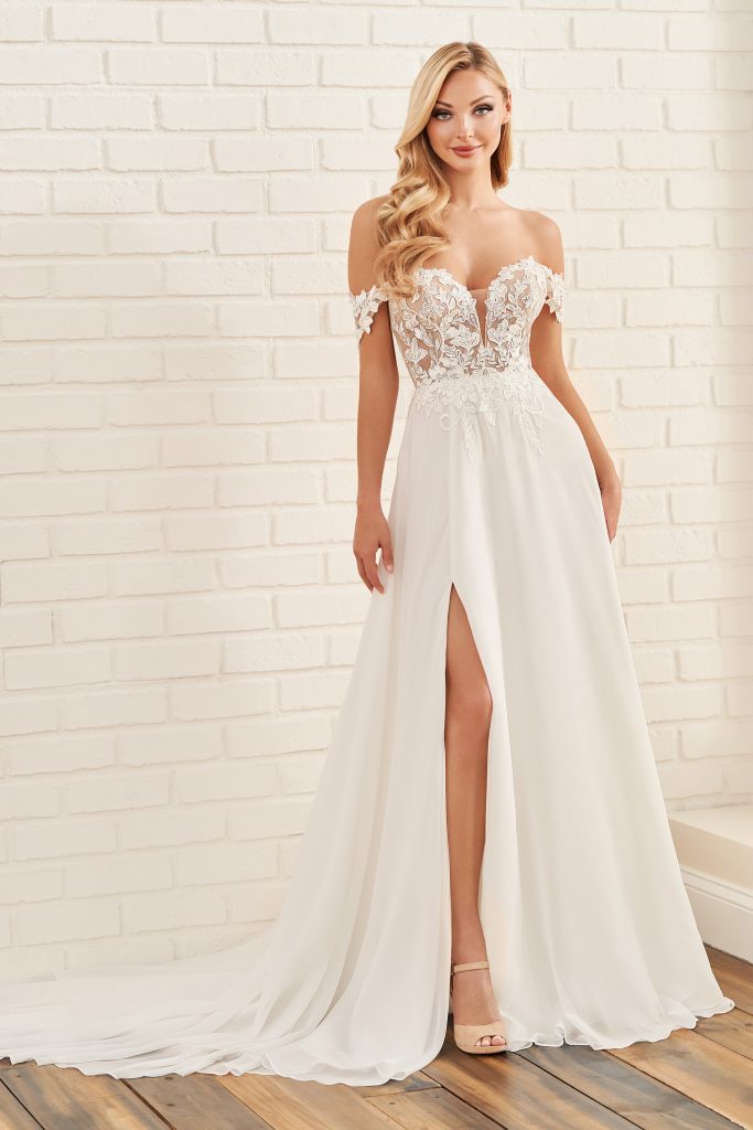 A smiling bride in front of a painted white brick wall, wearing a wedding dress with lace floral applique and a high slit.