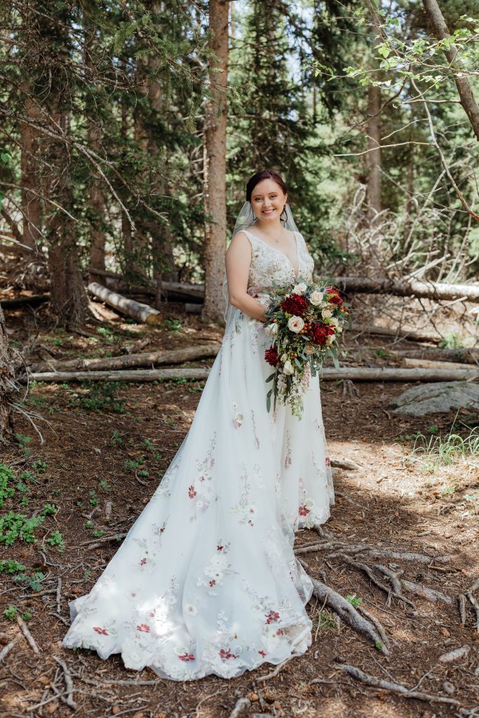 Bride standing in a wooded area wearing a floral appliqué gown and holding a rich, romantic bouquet.