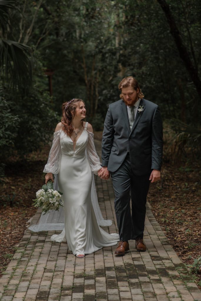 Bride and groom walking hand-in-hand down a brick path, surrounded by lush greenery.