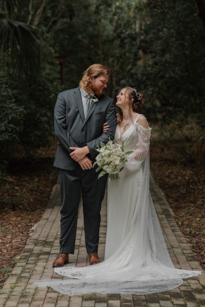 Bride and groom standing close on a forest path, bride holding a bouquet of white and green flowers.