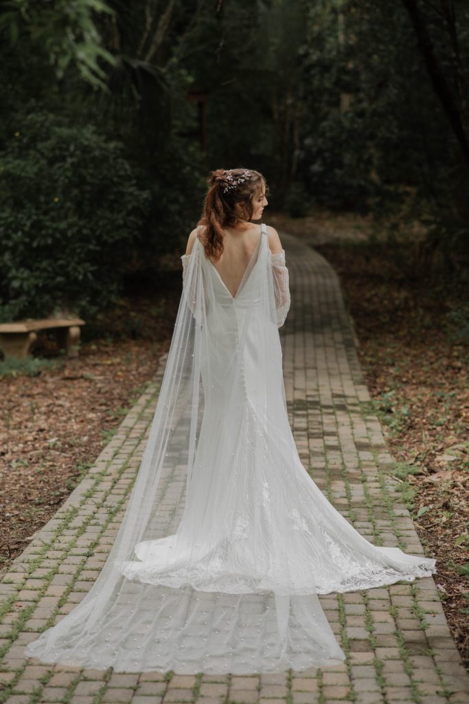 Bride walking alone on a forest path in a long white gown with draped off-shoulder sleeves.