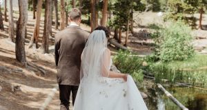 Bride and groom walk through a forested area beside a pond, seen from behind.