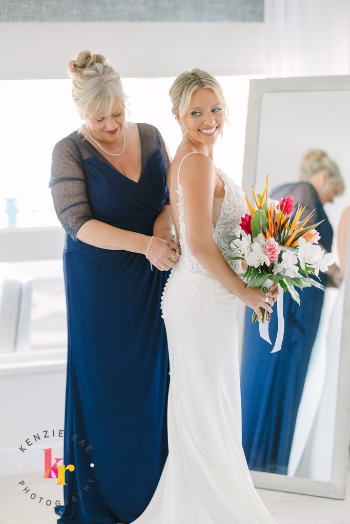 Mother helps bride button her dress as she holds a bouquet of colorful tropical flowers.