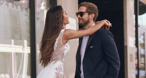 A bride in a fitted lace-back wedding gown embraces a groom in a navy suit and sunglasses on a city sidewalk, smiling at each other.