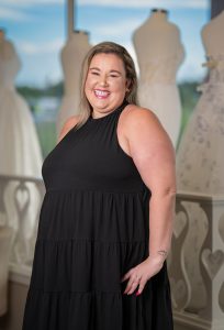 A woman in a long black dress is smiling with her pink lips while standing behind a display of wedding dresses.