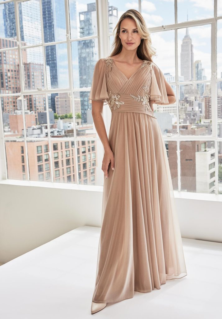 A woman stands near a city window wearing a soft taupe chiffon gown with flutter sleeves and floral appliqués on the shoulders and bodice.