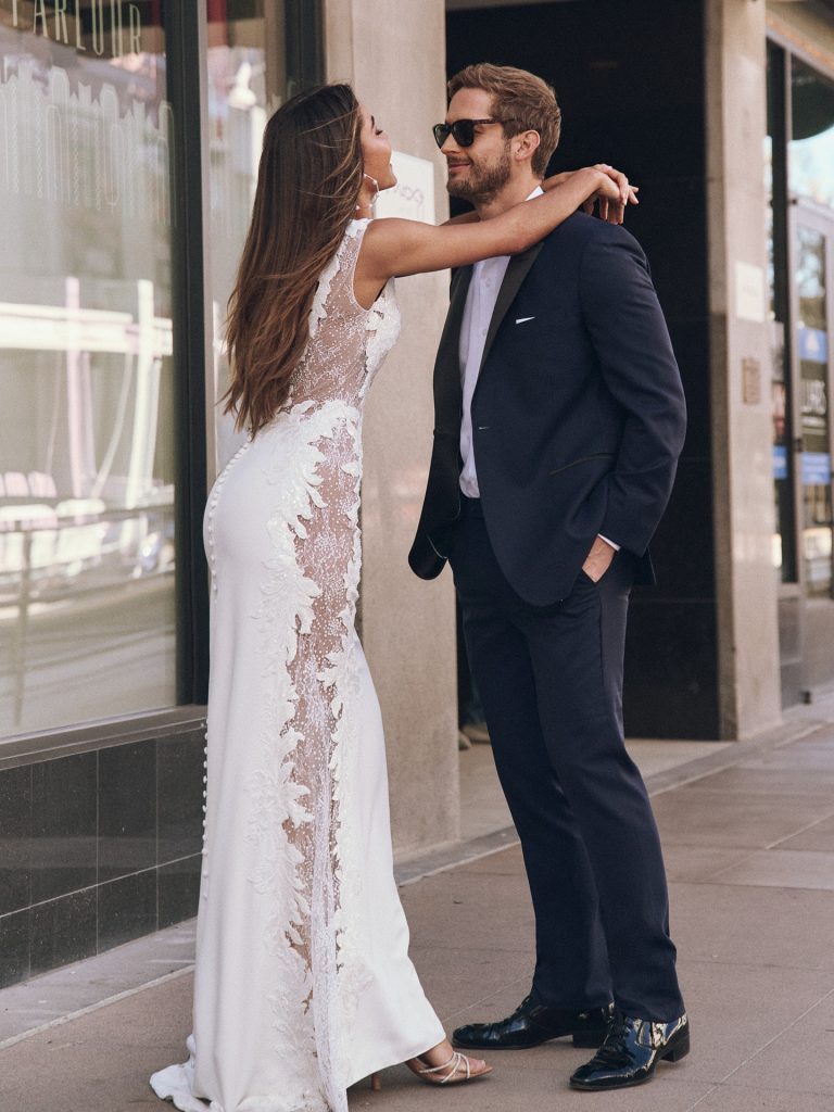 A bride and groom stand on a city sidewalk. The bride wears a sleek lace-back gown with a long train, while the groom wears a navy suit and sunglasses.