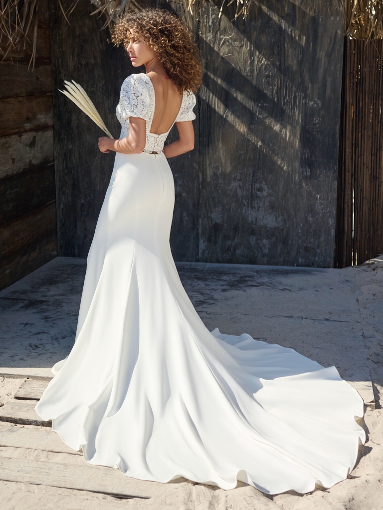 A bride poses on a sandy boardwalk wearing a crepe tropical wedding dress with lace puff sleeves and an open back.