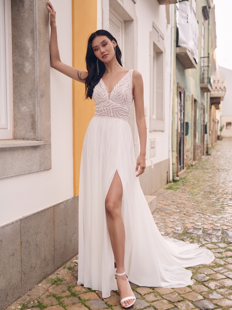 A bride leans against a wall in a boho lace gown with a plunging neckline, side slit, and illusion detailing, ideal for a tropical beach wedding.