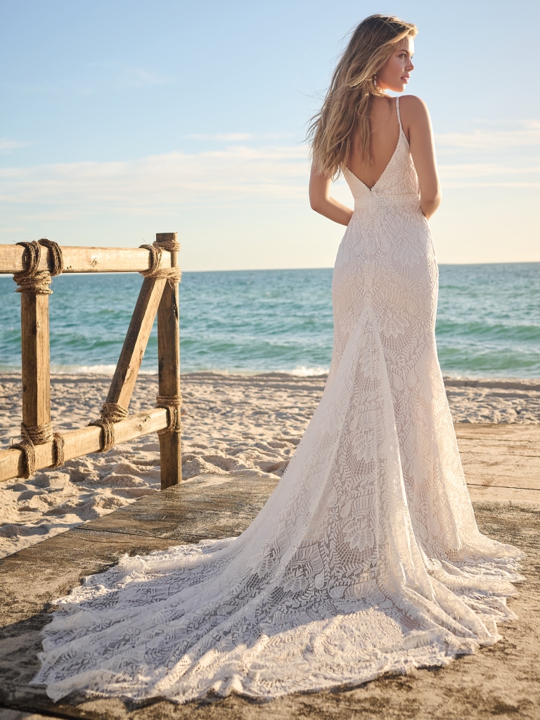 A bride gazes at the ocean while wearing a lace tropical wedding dress with thin straps and a full train, perfect for a seaside ceremony.