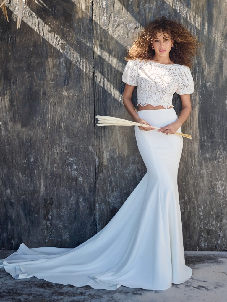 A bride holds palm fronds and wears a bohemian two-piece tropical wedding gown with a lace crop top and fitted skirt.