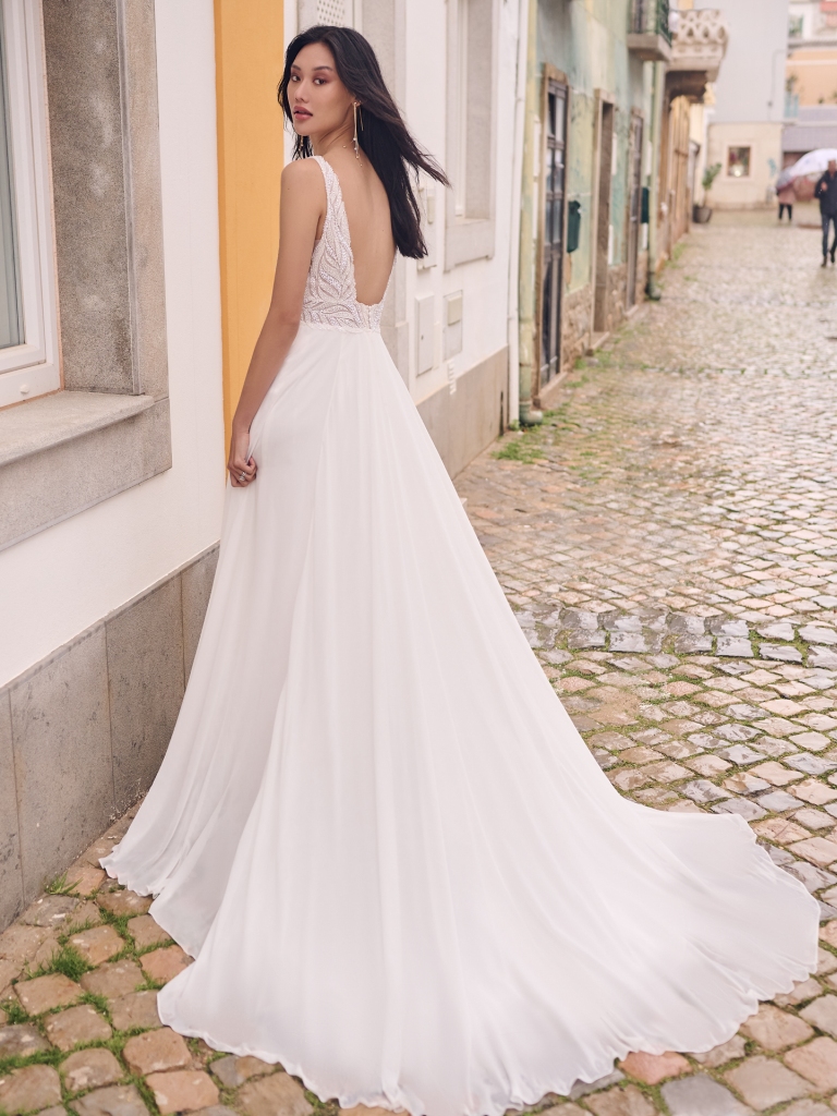 A bride stands on a cobblestone street in a sleeveless tropical wedding gown with a deep V-back and flowing chiffon train.