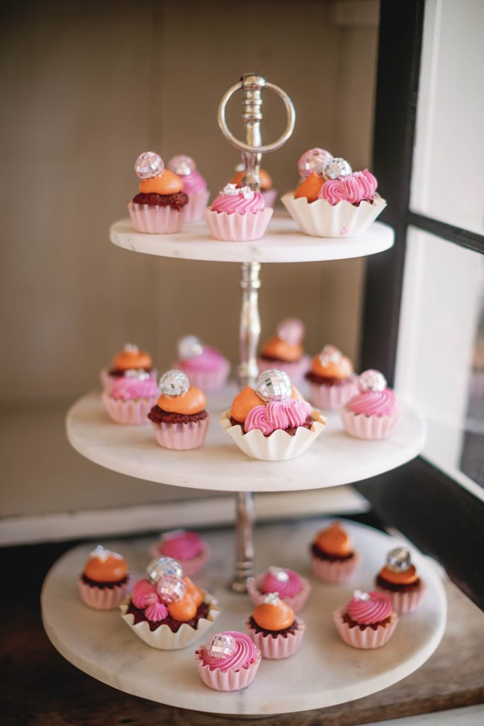 A close-up of a three-tiered dessert stand filled with cupcakes topped with pink frosting, orange cream, and miniature disco balls.