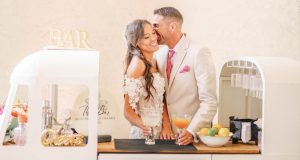 A bride and groom smile together while standing at a retro-style mobile bar, each holding a signature wedding cocktail garnished with citrus.