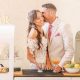 A bride and groom smile together while standing at a retro-style mobile bar, each holding a signature wedding cocktail garnished with citrus.
