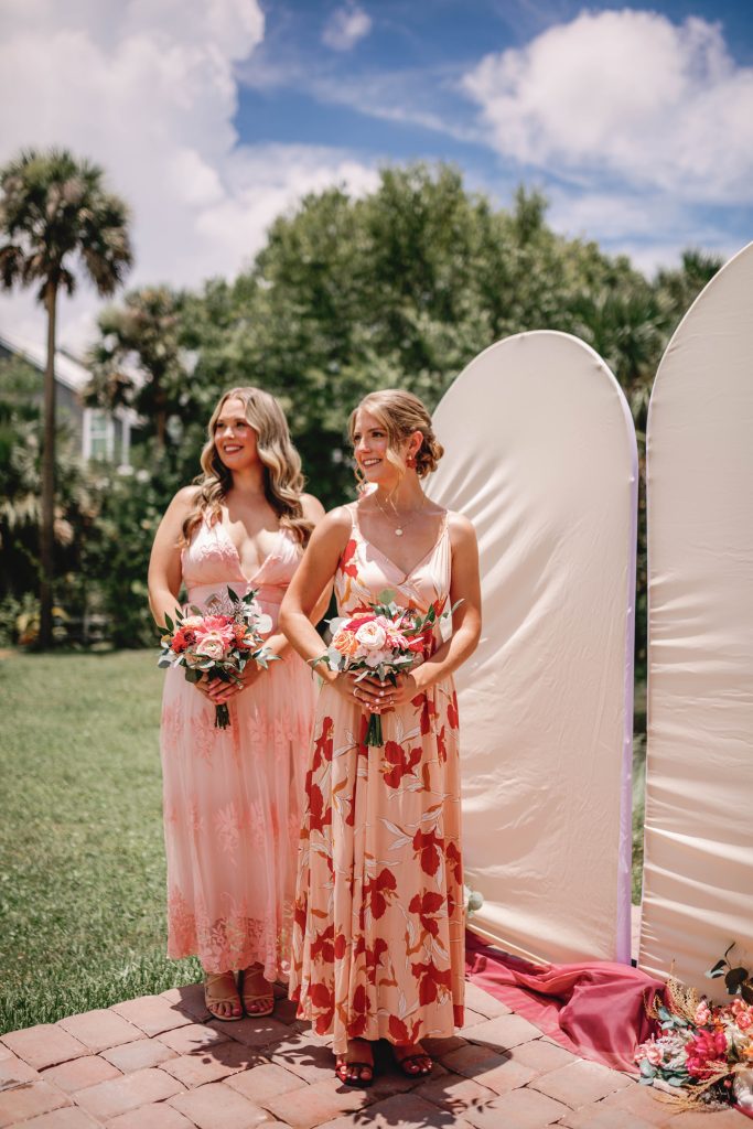 Two bridesmaids stand smiling during an outdoor wedding ceremony, each holding a bouquet of pink and coral flowers.
