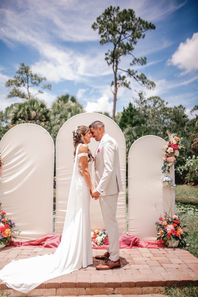 Bride and groom hold hands and smile under a blue sky during an outdoor ceremony with a unique arch.