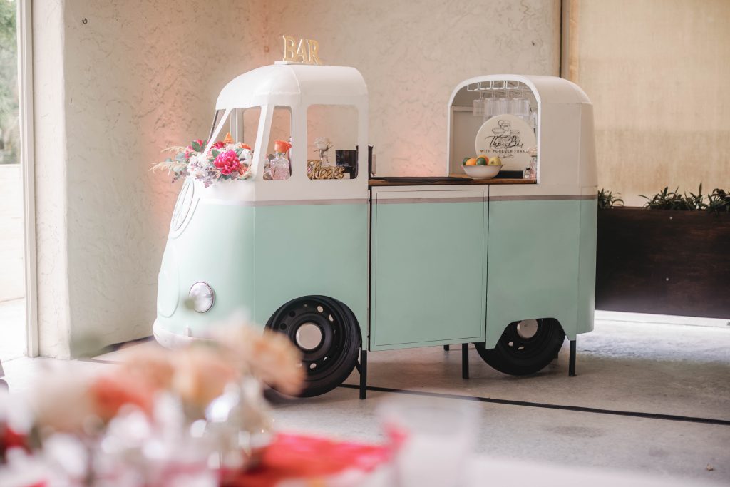 Mint green VW bus bar cart decorated with flowers and stocked with drinks, displayed at a wedding reception.