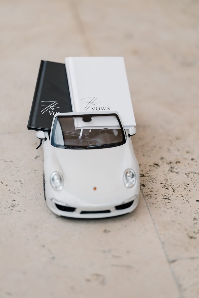Two wedding vow books placed inside a small white convertible model car on a tiled floor.