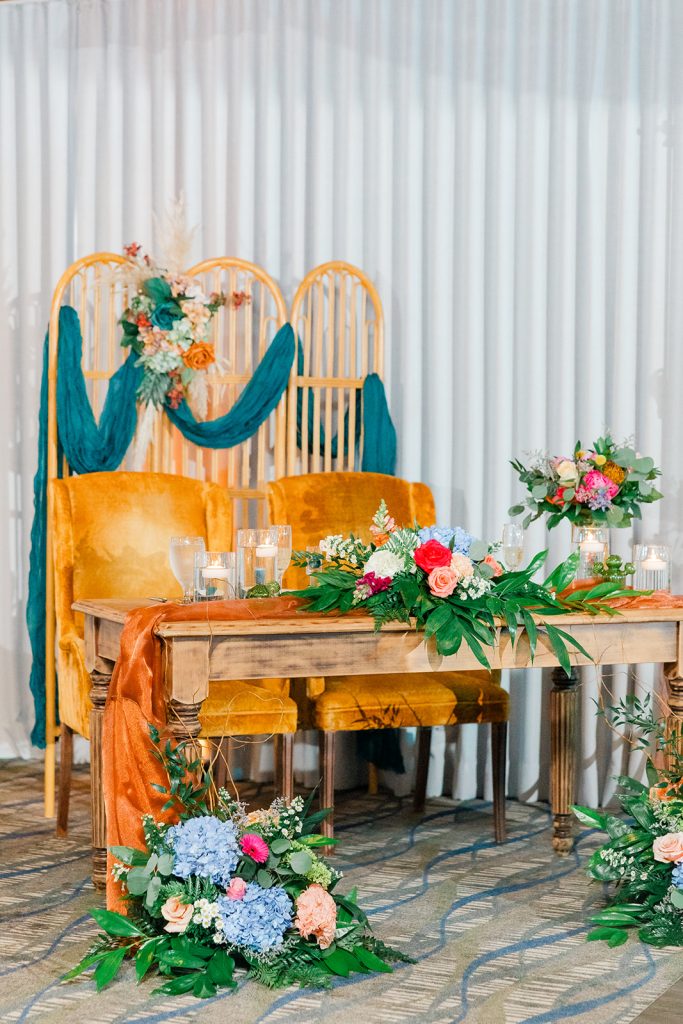 A sweetheart table with mustard yellow chairs, teal fabric accents, and colorful floral arrangements in a wedding reception space.