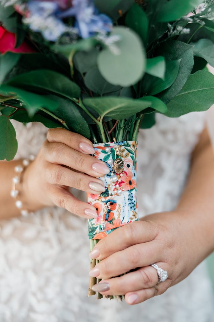 A close-up of a bride&rsquo;s hands holding a bouquet wrapped in floral fabric, adorned with small charms and jewelry.
