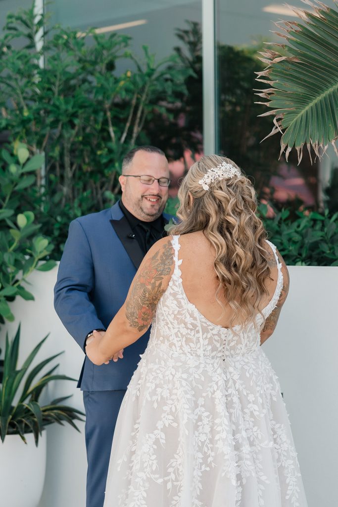 A groom smiling while holding hands with his bride, who is seen from the back in a lace gown, standing outdoors near greenery.