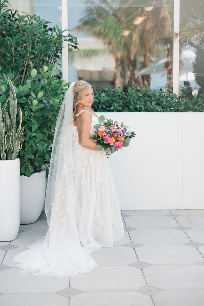A bride in a lace wedding gown holding a colorful bouquet, standing next to large potted plants outdoors.