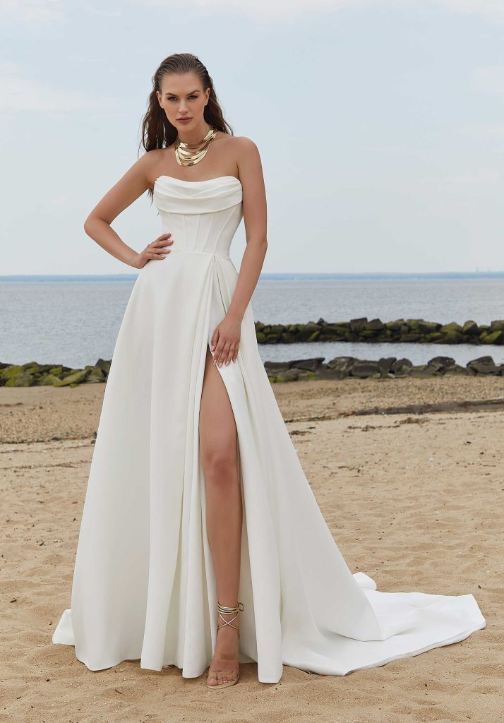 Bride in a strapless gown with a pleated bodice and high leg slit, wearing a gold necklace and standing on a sandy beach.