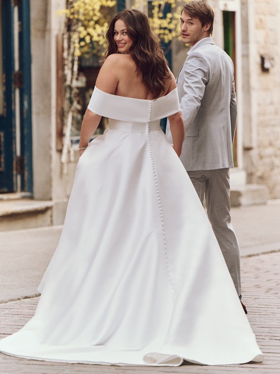 A smiling bride in an off-the-shoulder satin gown walks outdoors with a groom in a light gray suit.