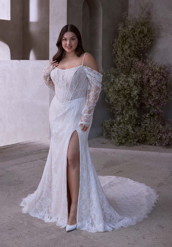 A bride in a lace gown with off-the-shoulder long sleeves and a high slit poses in front of textured stone walls.