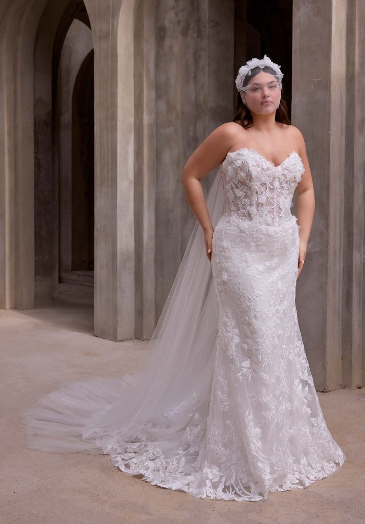 A bride wearing a strapless lace wedding gown with a floral headpiece and long train stands against tall stone arches.