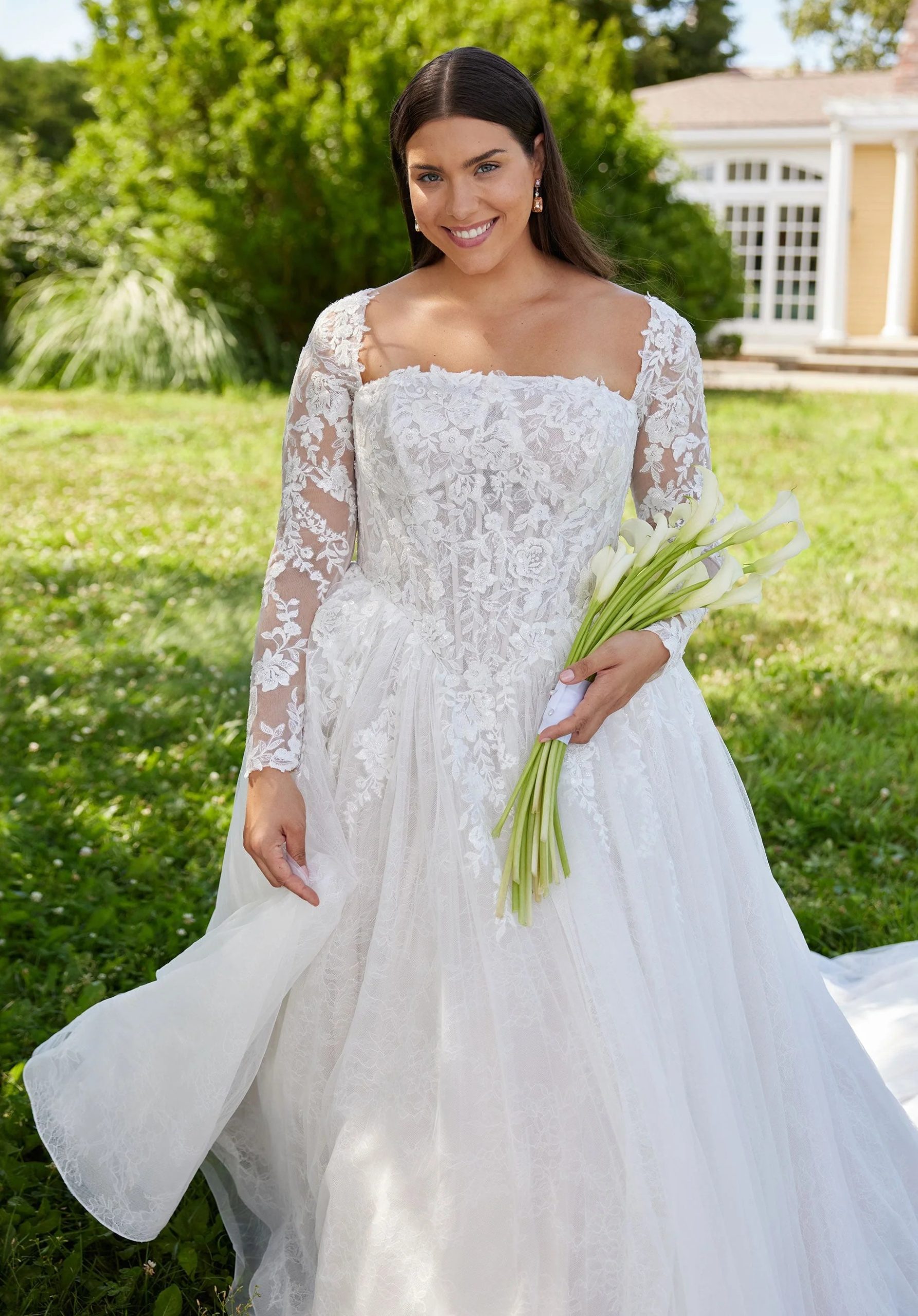 A smiling bride stands on grass wearing a long-sleeve lace ballgown and holding a bouquet of white calla lilies.