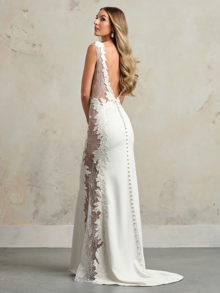 Bride in a fitted sleeveless gown with lace side panels and buttons down the back, standing against a textured neutral wall.