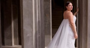 A bride looks back while standing between stone columns, wearing a strapless lace gown with a flowing tulle cape.
