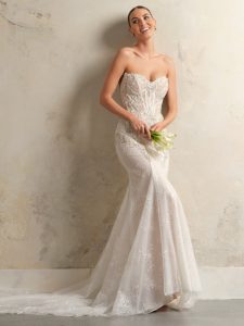 Bride in a strapless mermaid-style wedding dress holding a small bouquet, posing against a neutral wall.