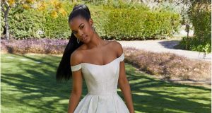 A bride stands outdoors in a garden wearing an off-the-shoulder white ball gown wedding dress with a fitted bodice and full skirt.