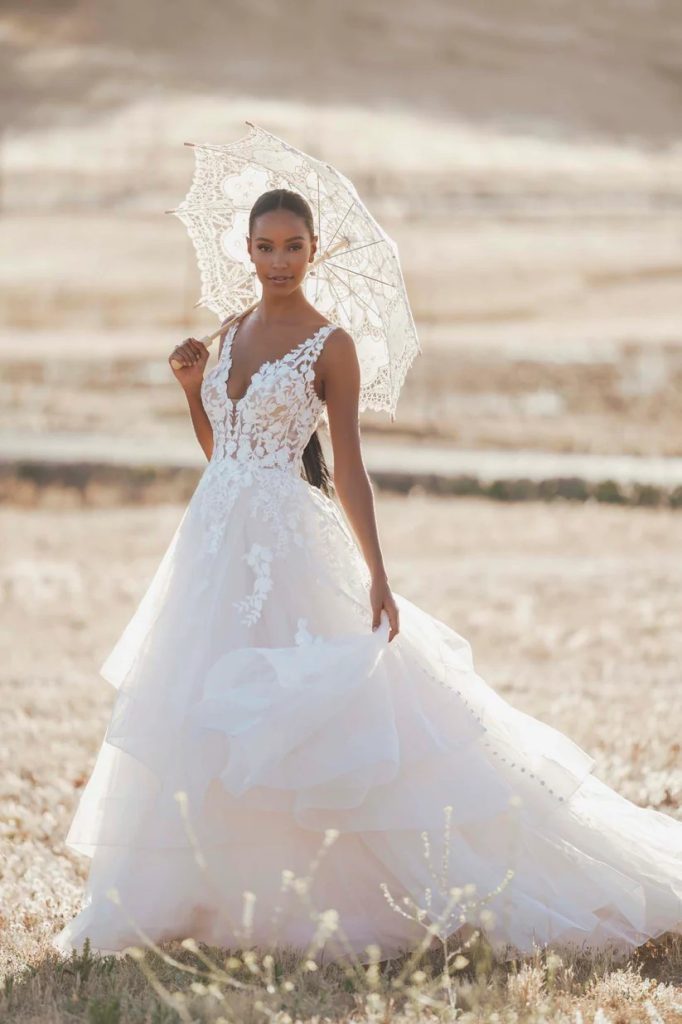 A bride wearing a sleeveless lace ball gown holds a white lace parasol while standing in a sunlit field.