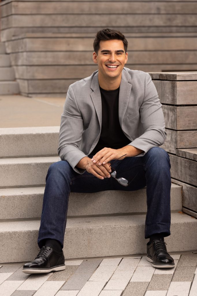 A man sits on outdoor steps wearing a gray blazer, black shirt, and jeans, smiling while holding eyeglasses in one hand.