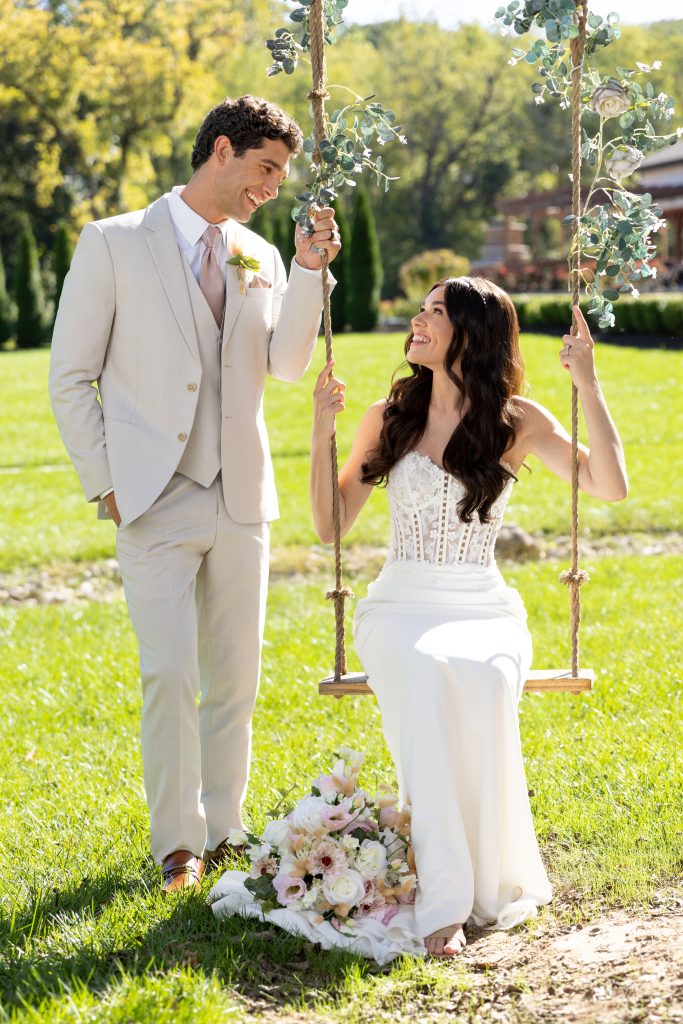 A groom in a light beige suit smiles at a bride in a white gown sitting on a swing decorated with greenery and flowers.