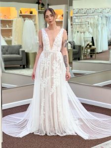 Bride wearing a lace A-line gown with a deep V neckline and flowing sheer train, standing inside a bridal boutique.
