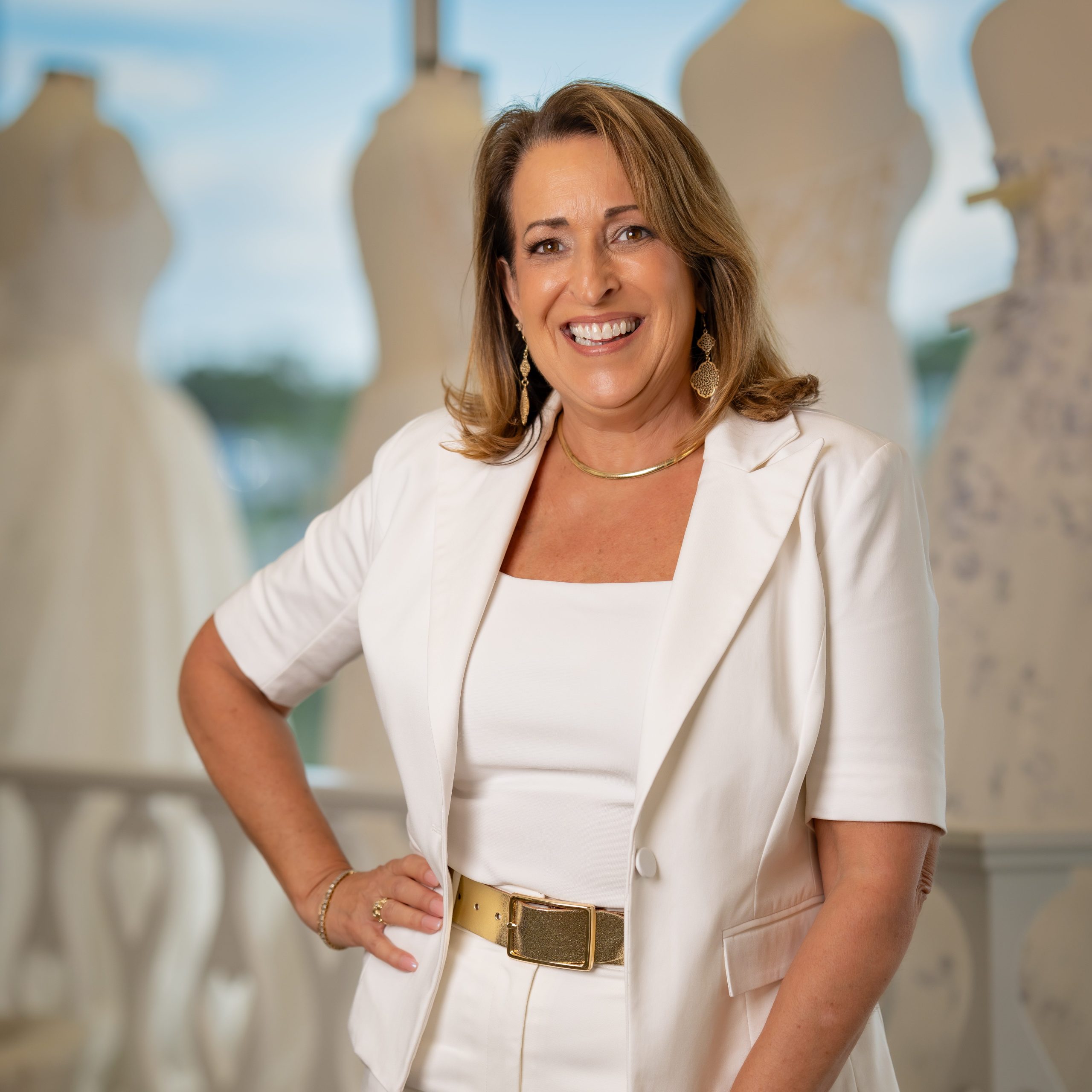 Cami Hester of Aurora Bridal is wearing a white top and bottom with a gold belt and gold accessories.