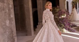 A bride wearing a high-neck lace long sleeve wedding dress with a full skirt, standing in a stone hallway with floral arrangements.