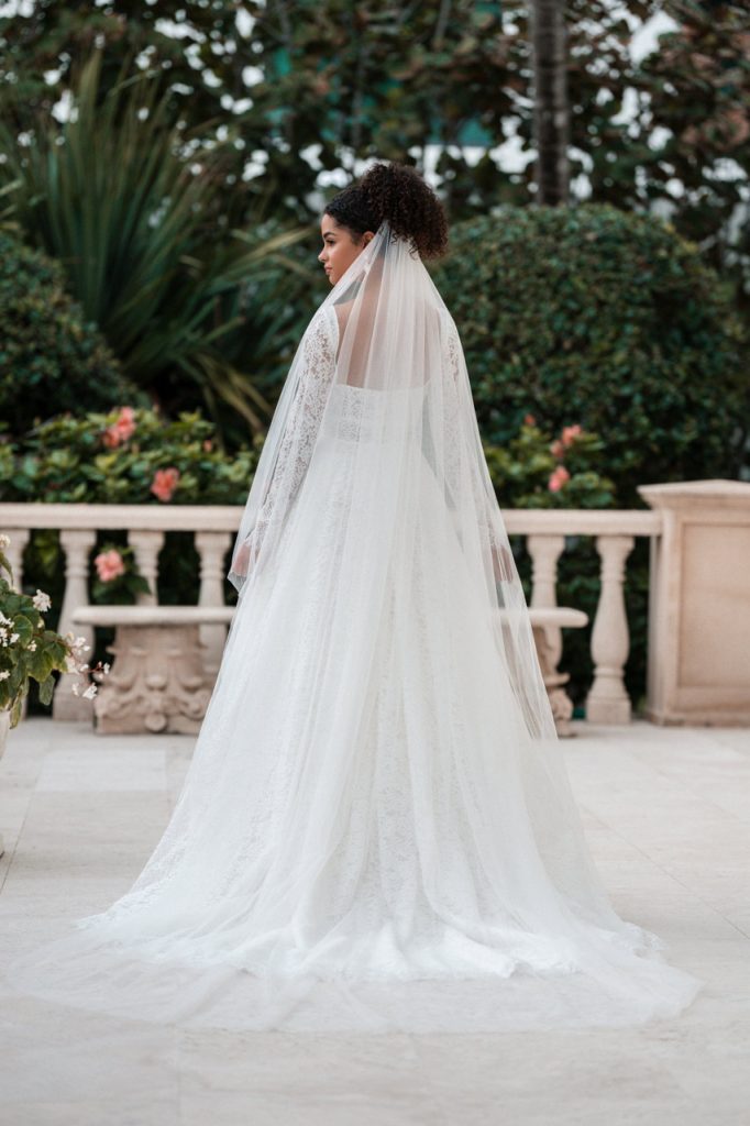 Back view of a bride in a lace long sleeve wedding dress with a flowing veil and full skirt, standing near stone balustrades and gardens.