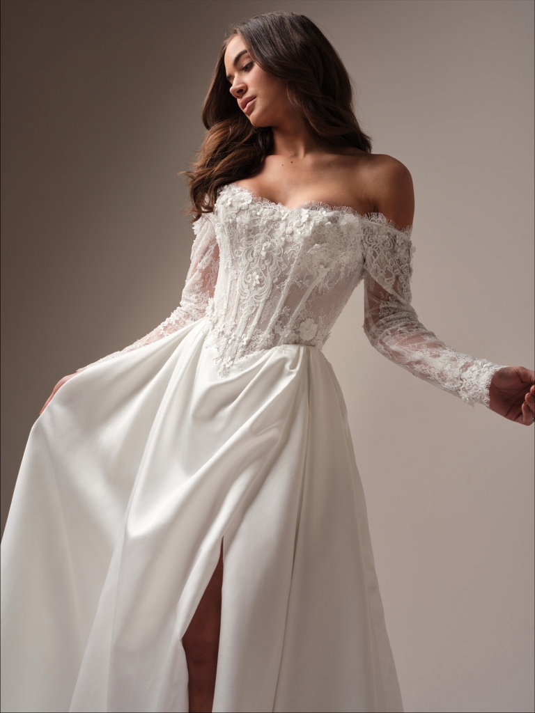 A bride in an off-the-shoulder lace long sleeve wedding dress with a high slit in the skirt, holding the gown fabric indoors.