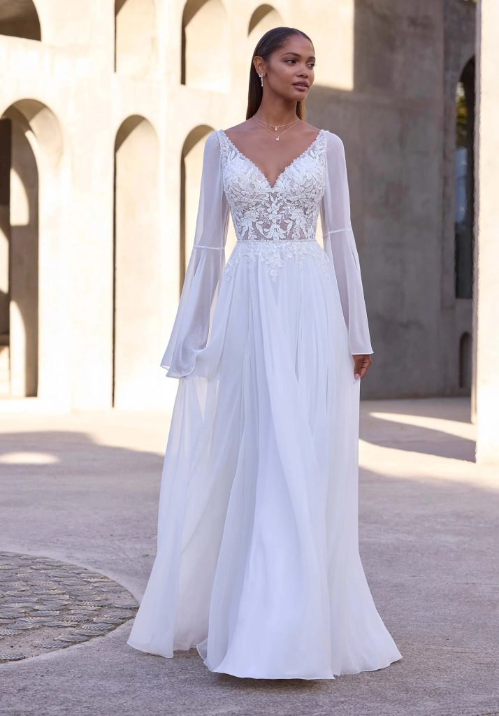 A bride in a flowing lace and chiffon long sleeve wedding dress with bell sleeves and a V-neckline, standing outside near stone arches.